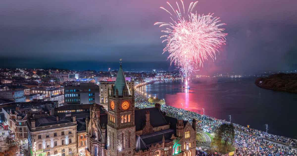 High-angle view of pink and white fireworks exploding over the River Foyle and the historic Guildhall during the Derry Halloween 40th Anniversary celebration in Derry, Northern Ireland.