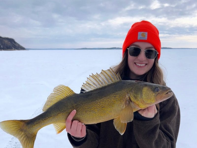 Taylor in an orange beanie and sunglasses holding a walleye she caught while ice fishing on a frozen lake.