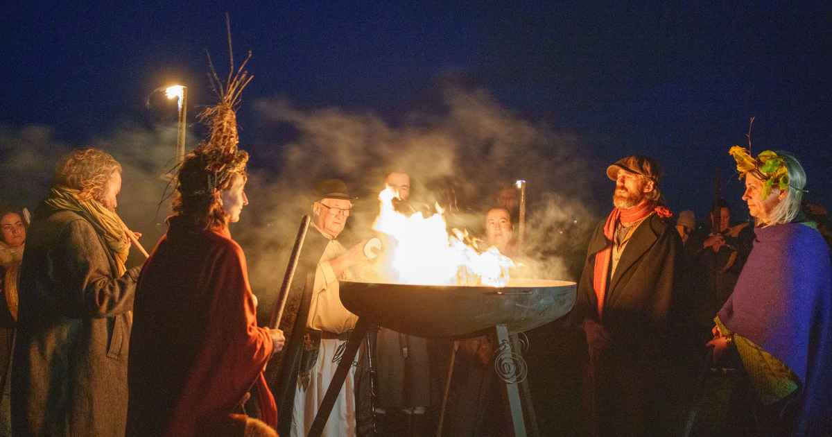 A group of people in traditional cloaks and headpieces gather around a large metal cauldron as a sacred Samhain fire is lit at the Púca Festival in County Meath, Ireland.
