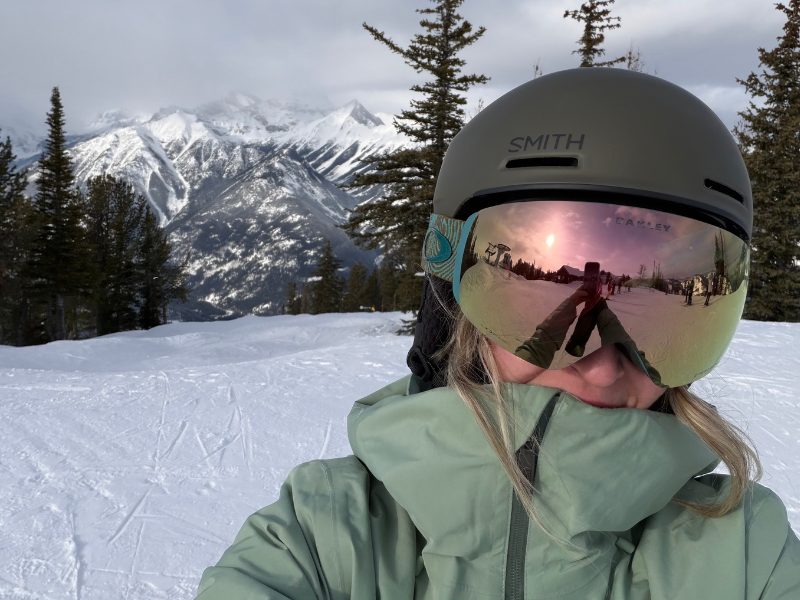 Taylor taking a selfie on the ski slopes in a green jacket and helmet, with snow-covered peaks and evergreen trees behind her.