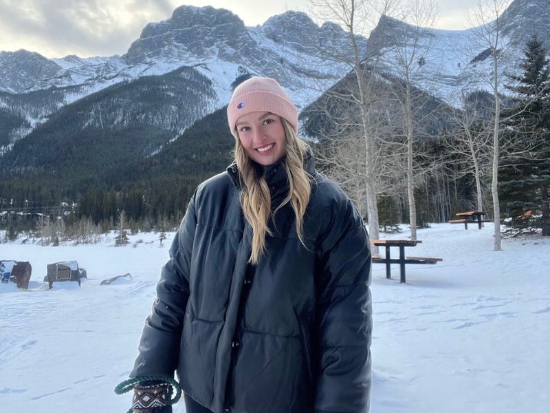 Taylor smiling in a pink beanie and dark puffer jacket, standing in front of snow-covered mountains near Canmore, Alberta.